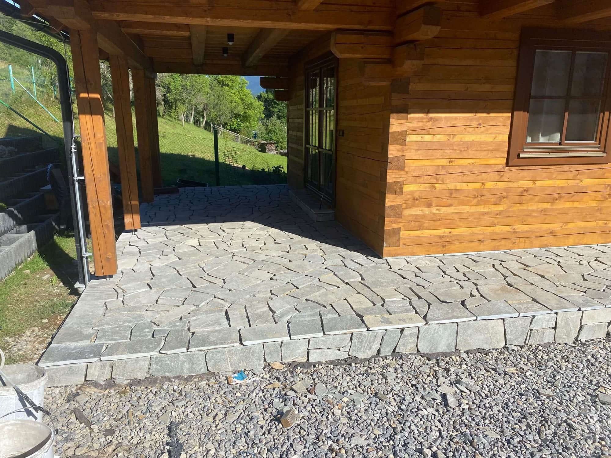 Kamenná dlažba pokračujúca z terasy pod drevené prestrešenie chaty